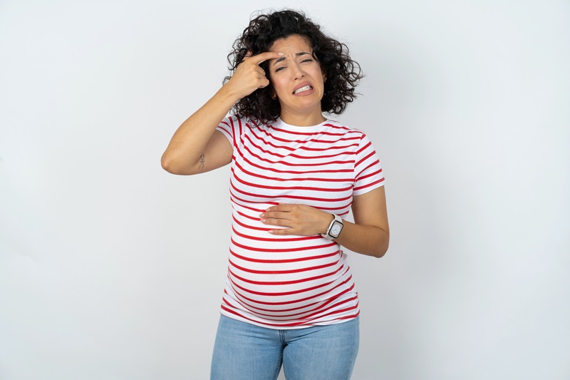 Osoba w ciąży dotykająca twarzy i brzucha na neutralnym tle, ilustrująca problem wyprysków na twarzy we wczesnej ciąży.